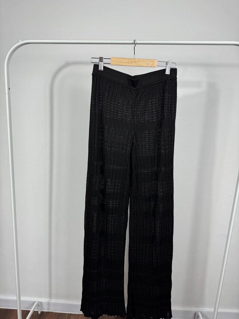 May include: Black, wide-leg pants with a textured, open weave design. The pants are hanging on a wooden hanger suspended from a white metal clothing rack. The fabric appears to be lightweight and suitable for warmer weather.