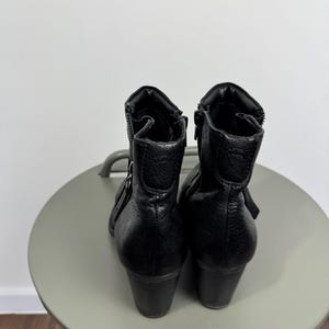 Pu&ograve; includere: Un paio di stivaletti in pelle nera con finitura strutturata. Gli stivaletti hanno una cerniera laterale, un piccolo dettaglio con fibbia e un tacco a blocco. Gli stivaletti sono esposti su una superficie rotonda grigio chiaro.