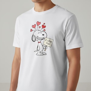 以下が含まれることがあります： 白いTシャツ。スヌーピーの漫画デザインで、「I LOVE YOU」と書かれた看板を持ち、頭の上に赤いハートが描かれています。デザインは黒、赤、白です。