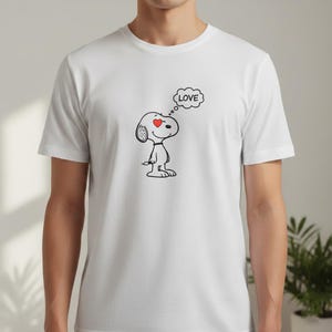 以下が含まれることがあります： 白のTシャツ。黒と白のスヌーピーの漫画のキャラクターが描かれており、赤いハートの目と「LOVE」という吹き出しがあります。このTシャツはクルーネックです。
