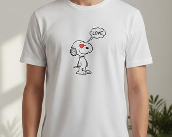 Könnte beinhalten: Weißes T-Shirt mit einem schwarz-weißen Snoopy-Cartoon. Snoopy hat ein rotes Herz als Auge und eine Sprechblase mit dem Wort "LOVE". Das T-Shirt hat einen Rundhalsausschnitt.