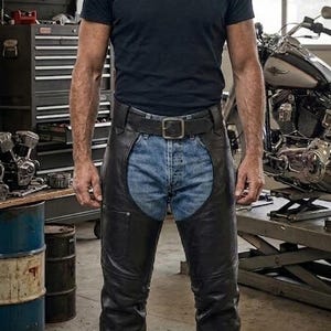 Op de afbeelding: Een donkerblauw T-shirt met korte mouwen gedragen met zwarte leren chaps over een lichtblauwe spijkerbroek. De chaps zijn voorzien van een riem aan de voorzijde met een zilverkleurige gesp, zijzakken en bedekken de volledige beenlengte.