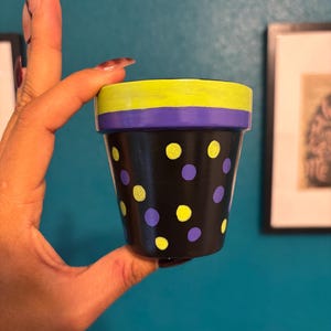 May include: A small, hand-painted black flower pot with yellow and purple accents. The pot features a pattern of yellow and purple polka dots. The rim is painted yellow and purple.