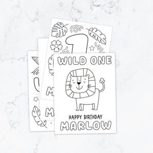 May include: A stack of white paper coloring sheets with black line art. The top sheet features a lion illustration, the text WILD ONE, HAPPY BIRTHDAY, and the name MARLOW. Other sheets show a large number one, tropical leaves, and flowers.