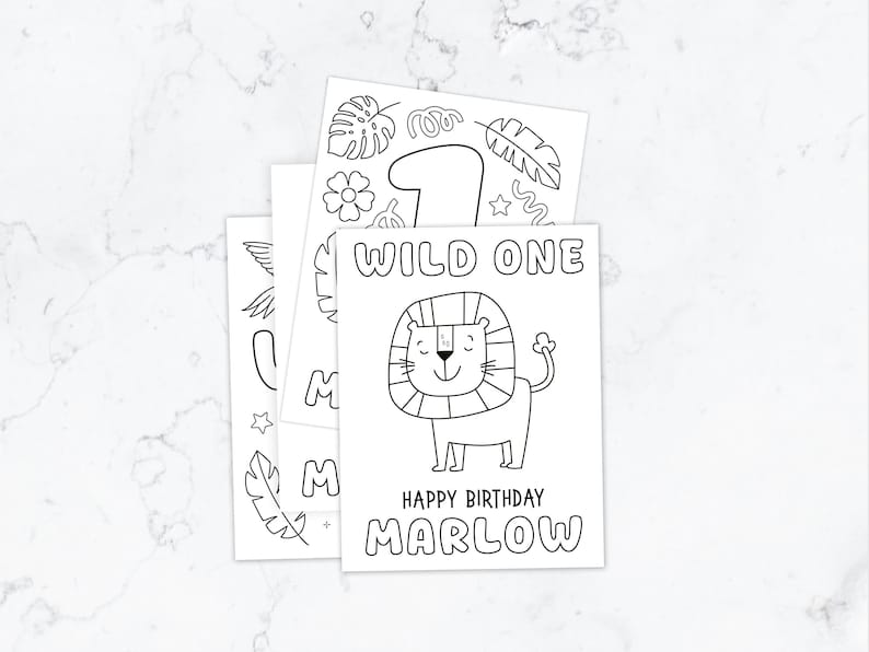 May include: A stack of white paper coloring sheets with black line art. The top sheet features a lion illustration, the text WILD ONE, HAPPY BIRTHDAY, and the name MARLOW. Other sheets show a large number one, tropical leaves, and flowers.