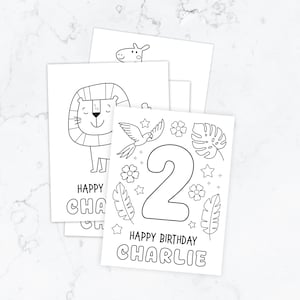 May include: A stack of white rectangular colouring pages featuring black line art. The top page displays a large number 2, tropical leaves, flowers, stars, and the text Happy Birthday Charlie. Other pages show a lion and a giraffe illustration.