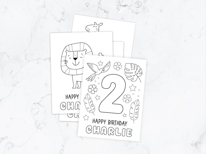 May include: A stack of white rectangular colouring pages featuring black line art. The top page displays a large number 2, tropical leaves, flowers, stars, and the text Happy Birthday Charlie. Other pages show a lion and a giraffe illustration.