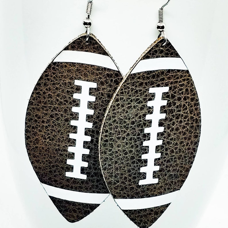 May include: A pair of brown leather earrings shaped like footballs with white stitching. The earrings have silver hooks.