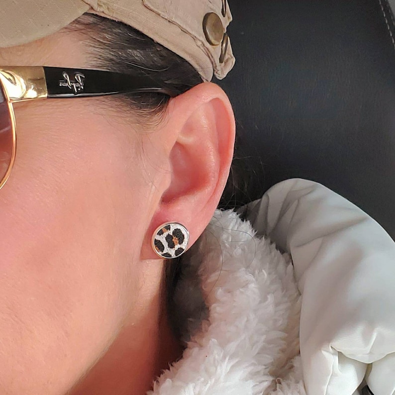 May include: A pair of small, round earrings with a leopard print design. The earrings are gold-toned and have a white background with black spots.