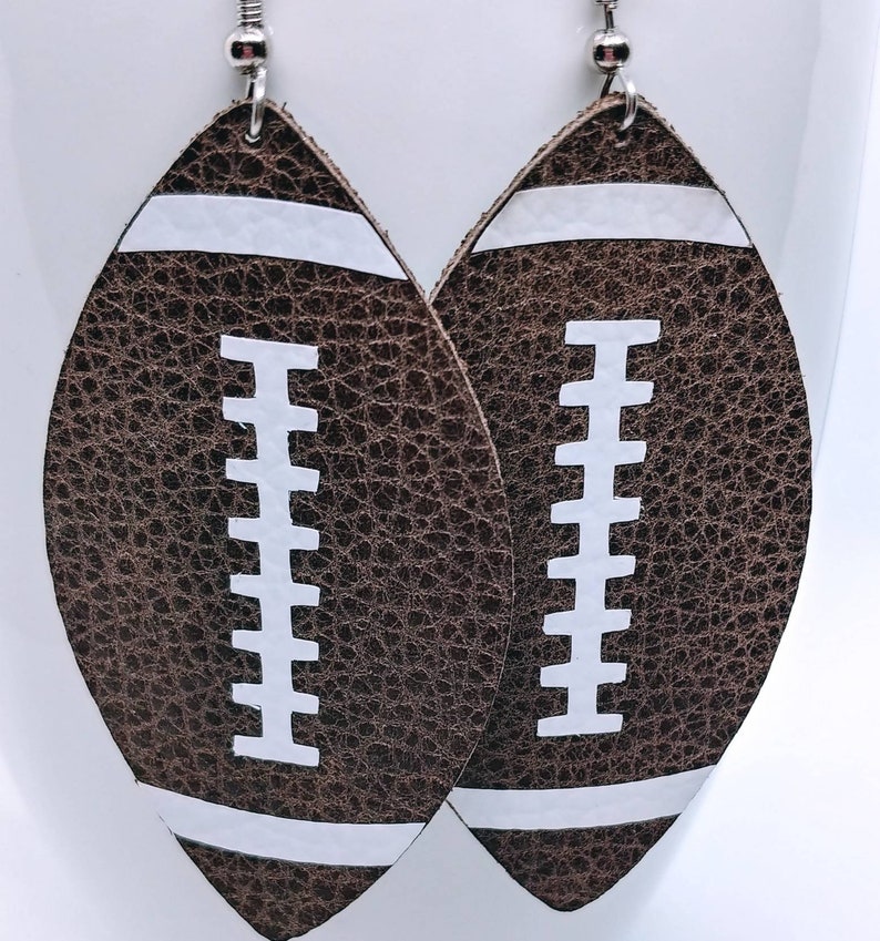 May include: A pair of brown leather earrings shaped like footballs with white stitching and white stripes.