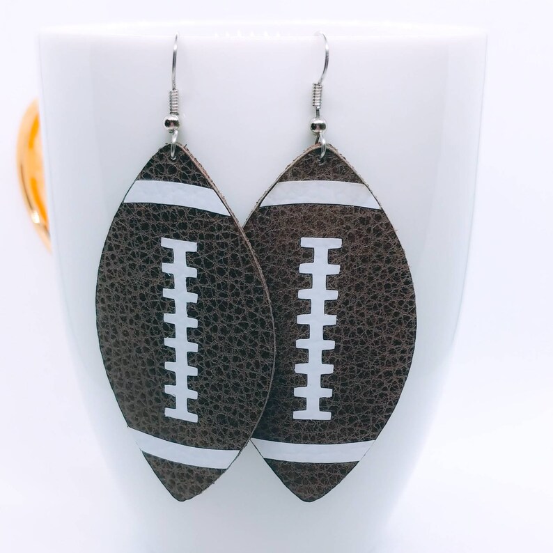 May include: A pair of brown leather earrings shaped like footballs with white stitching and white stripes.
