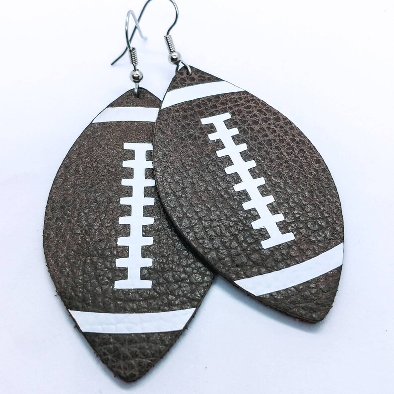 May include: A pair of brown leather earrings shaped like footballs with white stitching and white stripes.