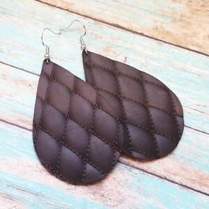 May include: A pair of brown leather teardrop earrings with a quilted pattern. The earrings have silver hooks.