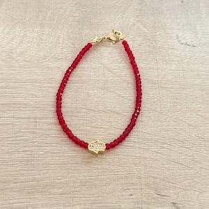 May include: Red beaded bracelet with a gold-colored Hamsa hand charm. The bracelet features small, round red beads strung together with a gold-colored clasp. The Hamsa hand charm is encrusted with small, clear crystals.