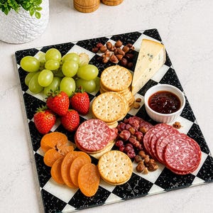 May include: A charcuterie board with a black and white checkered pattern. The board is filled with green grapes, red strawberries, crackers, salami, cheese, dried apricots, nuts, and a small bowl of jam.