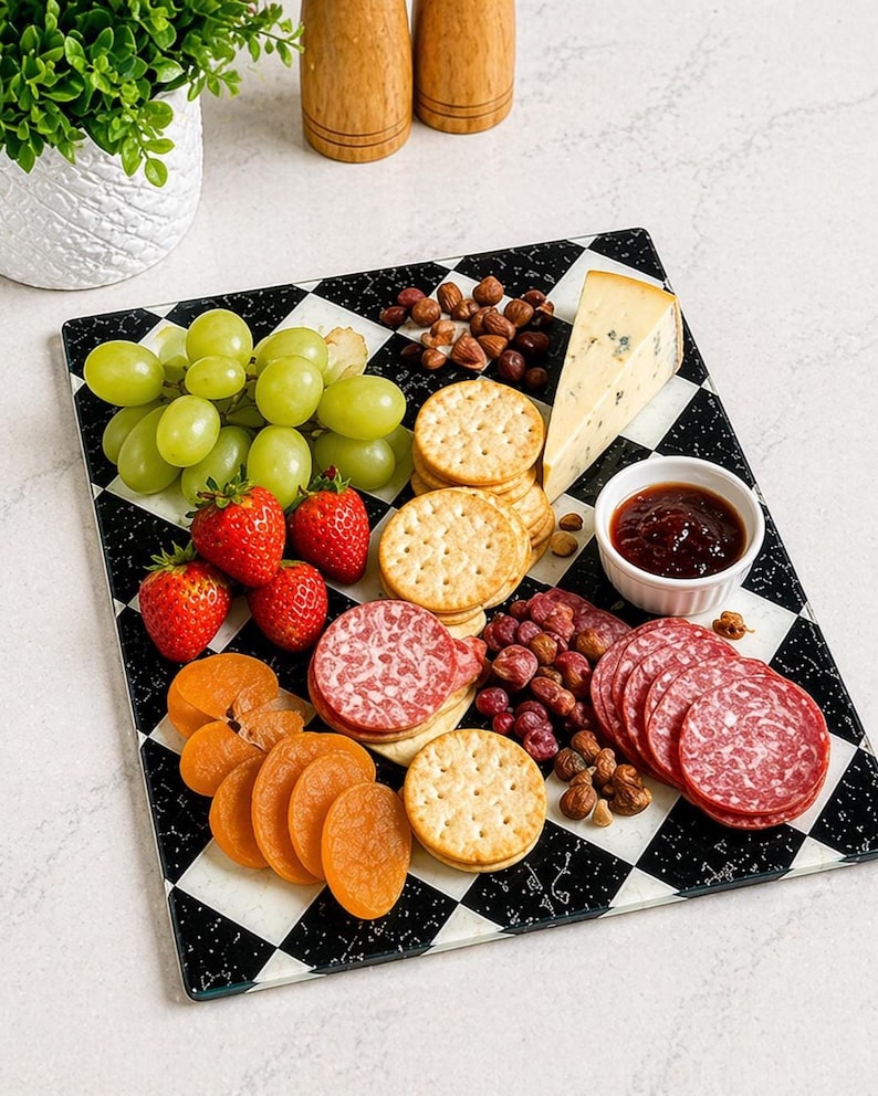 May include: A charcuterie board with a black and white checkered pattern. The board is filled with green grapes, red strawberries, crackers, salami, cheese, dried apricots, nuts, and a small bowl of jam.