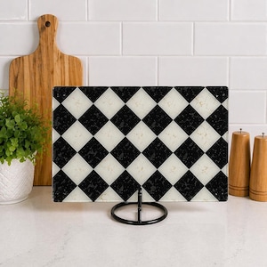 May include: A rectangular cutting board with a black and white checkered pattern, displayed on a black metal stand. The board is made of glass and has a textured surface. A wooden cutting board, a small potted plant, and salt and pepper shakers are in the background.
