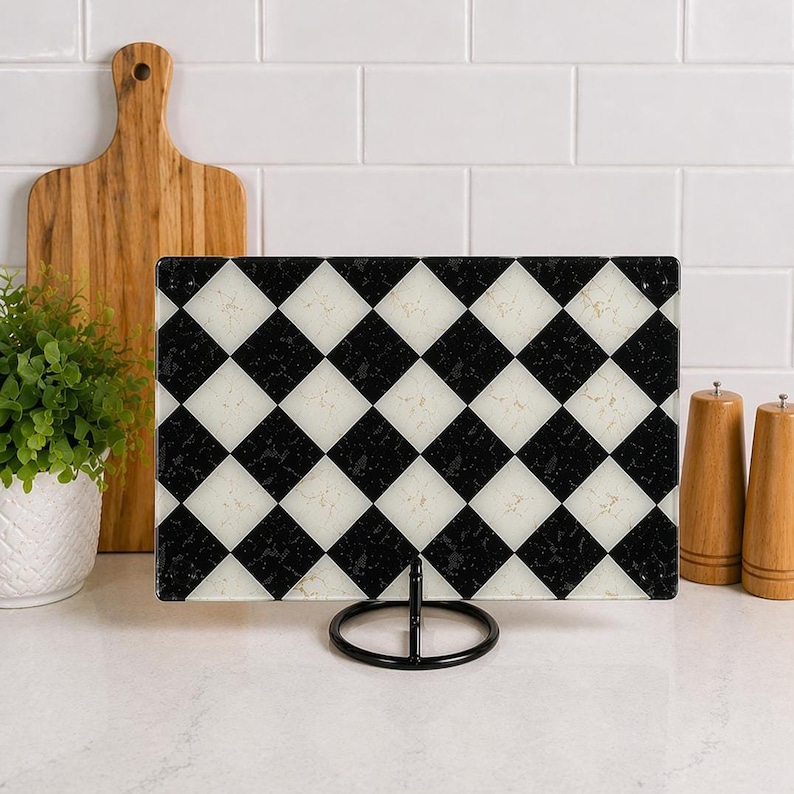 May include: A rectangular cutting board with a black and white checkered pattern, displayed on a black metal stand. The board is made of glass and has a textured surface. A wooden cutting board, a small potted plant, and salt and pepper shakers are in the background.