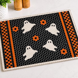 May include: A rectangular mosaic mat featuring a black hexagonal tile background with four white ghost figures and three orange flower shapes. The design is framed by a border of alternating orange and black square tiles with a white outer edge.