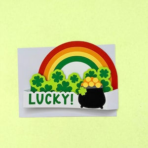 May include: A St. Patrick's Day greeting card with a rainbow, green shamrocks, and a pot of gold. The word "LUCKY!" is printed in green. The card is on a light yellow background.