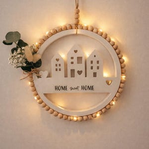 May include: A circular wooden wall decoration with a string of lights and wooden beads. It features three house cutouts and the text "HOME sweet HOME". A small floral arrangement is attached to the side.