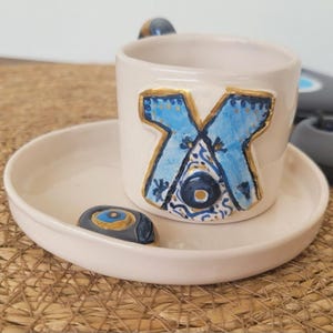 May include: A white ceramic coffee cup and saucer set. The cup features a blue and gold design with an eye motif. A small, decorative stone with an eye design sits on the saucer. The set is on a woven surface.