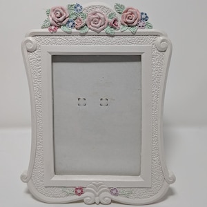 Op de afbeelding: Witte rechthoekige fotolijst met een bloemenontwerp. Het frame is versierd met roze rozen, blauwe en roze bloemen aan de boven- en onderkant. Het frame heeft een decoratieve, getextureerde rand.