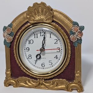 May include: A mantel clock with a gold-colored frame, a deep burgundy textured background, and a white face featuring black Arabic numerals. The clock is decorated with sculpted floral accents on both sides and a shell motif at the top.