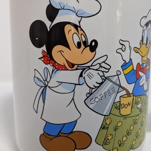 May include: White ceramic coffee mug featuring Mickey Mouse in a chef's outfit pouring coffee from a silver pot. Donald Duck is also present. The mug has a green floral tablecloth design and the word "COFFEE" on the pot.