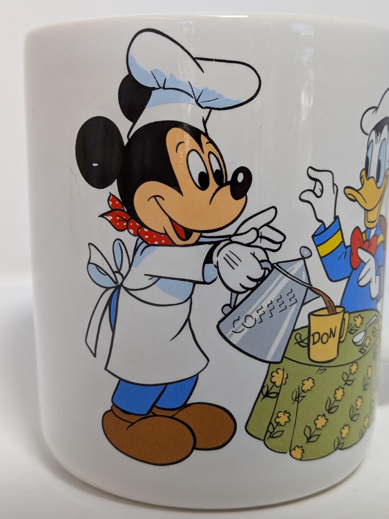 May include: White ceramic coffee mug featuring Mickey Mouse in a chef's outfit pouring coffee from a silver pot. Donald Duck is also present. The mug has a green floral tablecloth design and the word "COFFEE" on the pot.