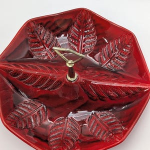May include: A glossy red ceramic serving dish with a divided interior and a central metal handle. The surface features a raised, embossed leaf pattern throughout the bowl, with darker red accents highlighting the texture of the leaves.