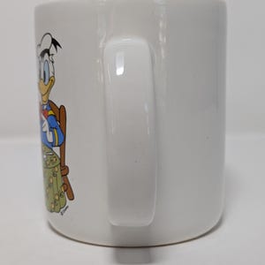 May include: White ceramic mug featuring a colorful illustration of Donald Duck in a dining setting. The handle is a solid white, and the mug has a glossy finish. The design includes a table and chair.