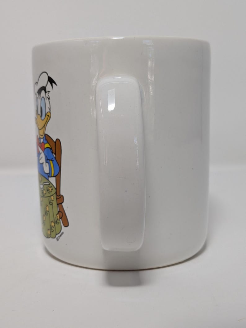 May include: White ceramic mug featuring a colorful illustration of Donald Duck in a dining setting. The handle is a solid white, and the mug has a glossy finish. The design includes a table and chair.