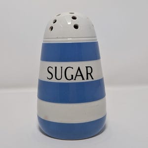 May include: A ceramic sugar shaker with a white top and a blue and white striped body. The word "SUGAR" is printed in black on the front. The shaker has a series of small holes on the top for dispensing sugar.