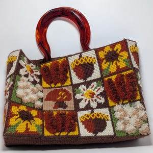 May include: A rectangular handbag crafted from needlepoint canvas featuring a grid of squares with embroidered motifs of sunflowers, acorns, mushrooms, and textured floral patterns. The bag includes a rounded, translucent amber-colored handle.