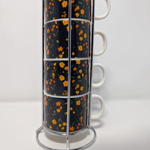 May include: A set of four stacked coffee mugs with a floral pattern in yellow and orange on a black background. The mugs have white handles and are held in a chrome metal rack.
