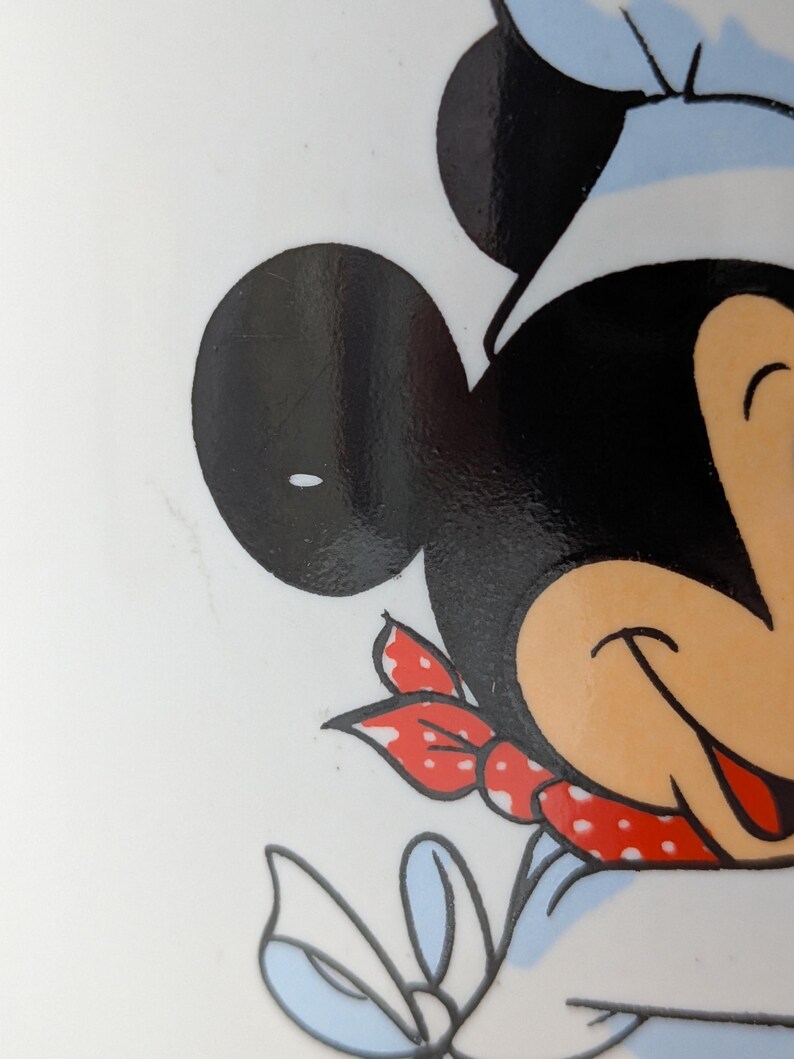 May include: Close-up of a cartoon character's face, featuring large black ears, a chef's hat, and a red polka-dot neckerchief. The character's face is a light tan color, with a smiling expression. The background is white.