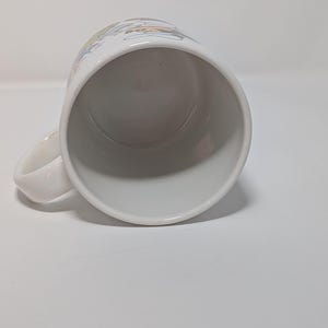 May include: White ceramic mug with a curved handle. The mug has a colorful design around the top rim. The interior of the mug is white. The mug is set against a white background.