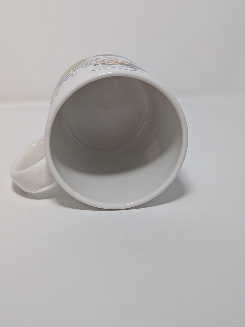 May include: White ceramic mug with a curved handle. The mug has a colorful design around the top rim. The interior of the mug is white. The mug is set against a white background.
