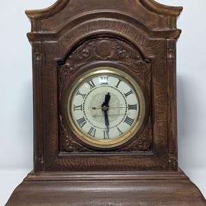 May include: A brown wooden mantel clock with a gold-tone clock face and Roman numerals. The clock has an ornate design with decorative carvings and a curved top. The clock face is cream-colored with black hands.