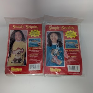 May include: Two Simply Sequins craft kits in clear plastic packaging. The left kit features a yellow shirt with a mouse design, while the right kit shows a blue shirt with a stocking design. Both red-bordered packages include iron-on charts.