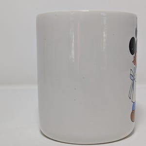 May include: White ceramic mug featuring a cartoon character design. The mug has a cylindrical shape with a rounded base. The cartoon character has black ears, a red bow, and a blue outfit. The mug is suitable for hot or cold beverages.