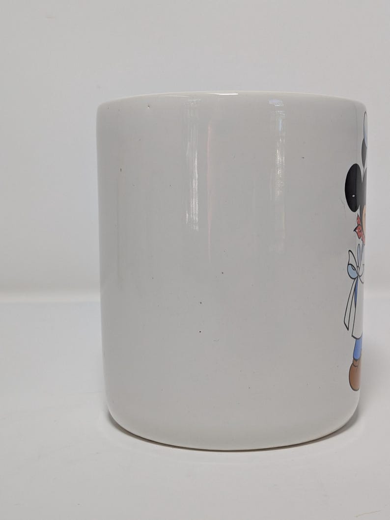 May include: White ceramic mug featuring a cartoon character design. The mug has a cylindrical shape with a rounded base. The cartoon character has black ears, a red bow, and a blue outfit. The mug is suitable for hot or cold beverages.