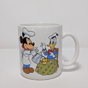 May include: White ceramic coffee mug featuring Mickey Mouse and Donald Duck. Mickey, in a chef's hat, pours coffee for Donald, who is seated at a table. The mug has a handle and a colorful illustration of the Disney characters.