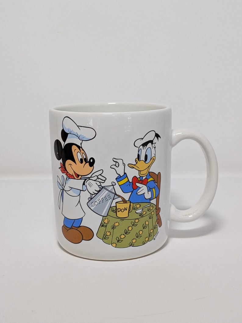 May include: White ceramic coffee mug featuring Mickey Mouse and Donald Duck. Mickey, in a chef's hat, pours coffee for Donald, who is seated at a table. The mug has a handle and a colorful illustration of the Disney characters.
