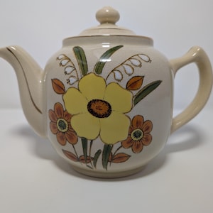 May include: Cream-colored ceramic teapot with a lid and handle. The teapot is decorated with a large yellow flower, smaller orange flowers, and green leaves. The spout and handle are a matching cream color.