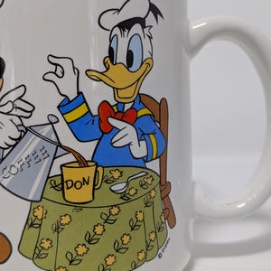 May include: White ceramic mug featuring Donald Duck pouring coffee into a yellow mug labeled "DON". Donald Duck is wearing a blue jacket, red bow tie, and chef's hat. The table has a green floral tablecloth. The mug has a white handle.