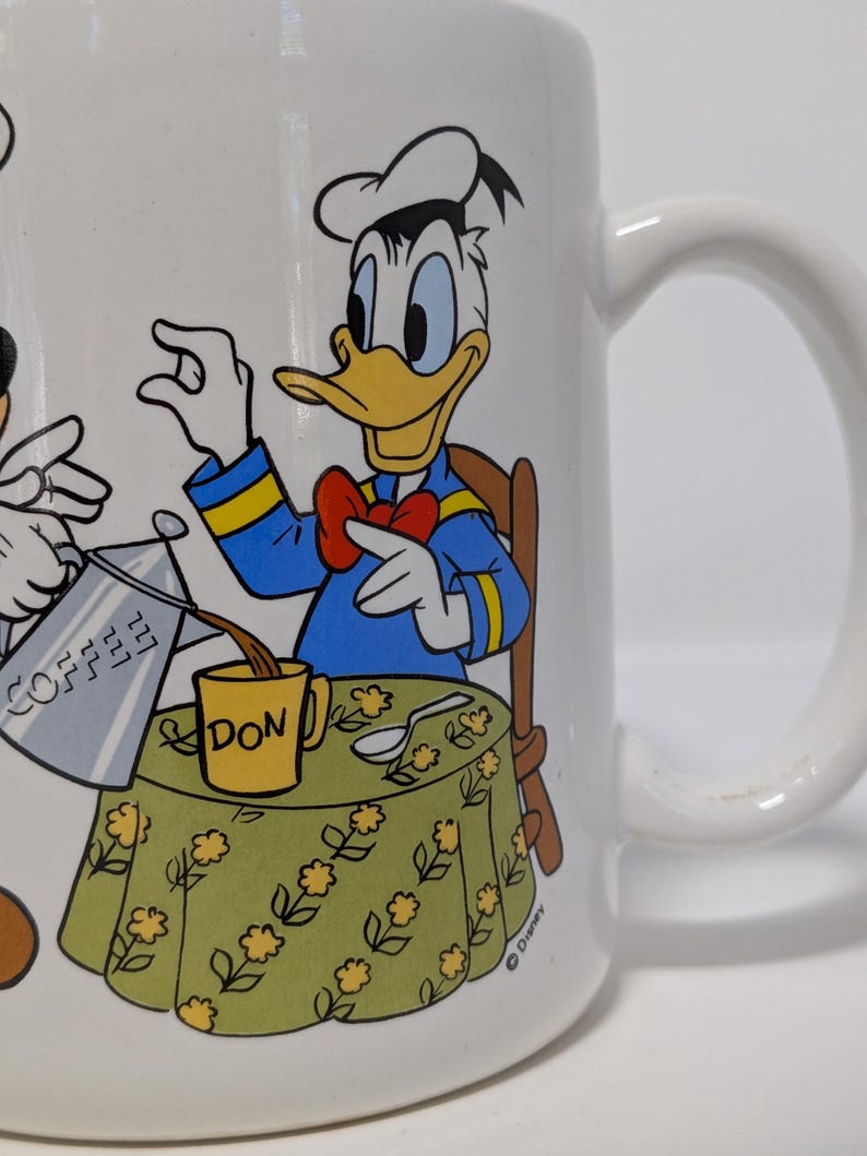 May include: White ceramic mug featuring Donald Duck pouring coffee into a yellow mug labeled "DON". Donald Duck is wearing a blue jacket, red bow tie, and chef's hat. The table has a green floral tablecloth. The mug has a white handle.