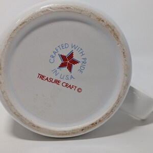May include: White ceramic mug with a handle, viewed from the bottom. The base features the text "Crafted with Pride in U.S.A." and "Treasure Craft" in red and blue. The mug has a clean, simple design.