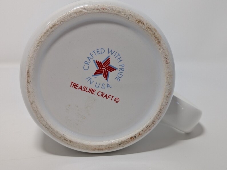 May include: White ceramic mug with a handle, viewed from the bottom. The base features the text "Crafted with Pride in U.S.A." and "Treasure Craft" in red and blue. The mug has a clean, simple design.